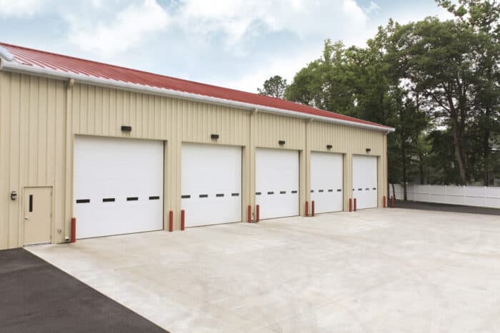 Four large white garage doors on a beige building with a red roof; paved area in front and trees in the background.