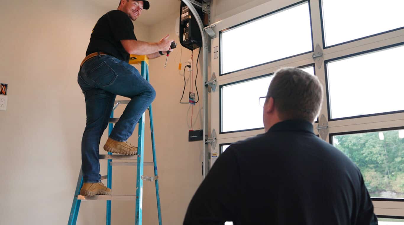 In a busy garage, one person stands on a ladder expertly working on electrical equipment for the commercial overhead doors, while the other carefully observes from the ground.