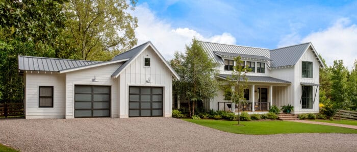 Modern white farmhouse with metal roof, two garage doors, large windows, and a landscaped front yard. Trees surround the house under a blue sky.