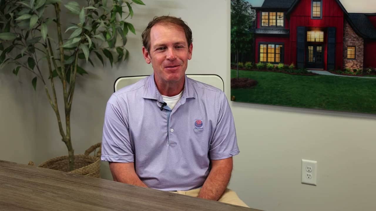 A man in a light purple polo shirt sits at a desk, smiling, with a plant and a framed picture of a house in the background.