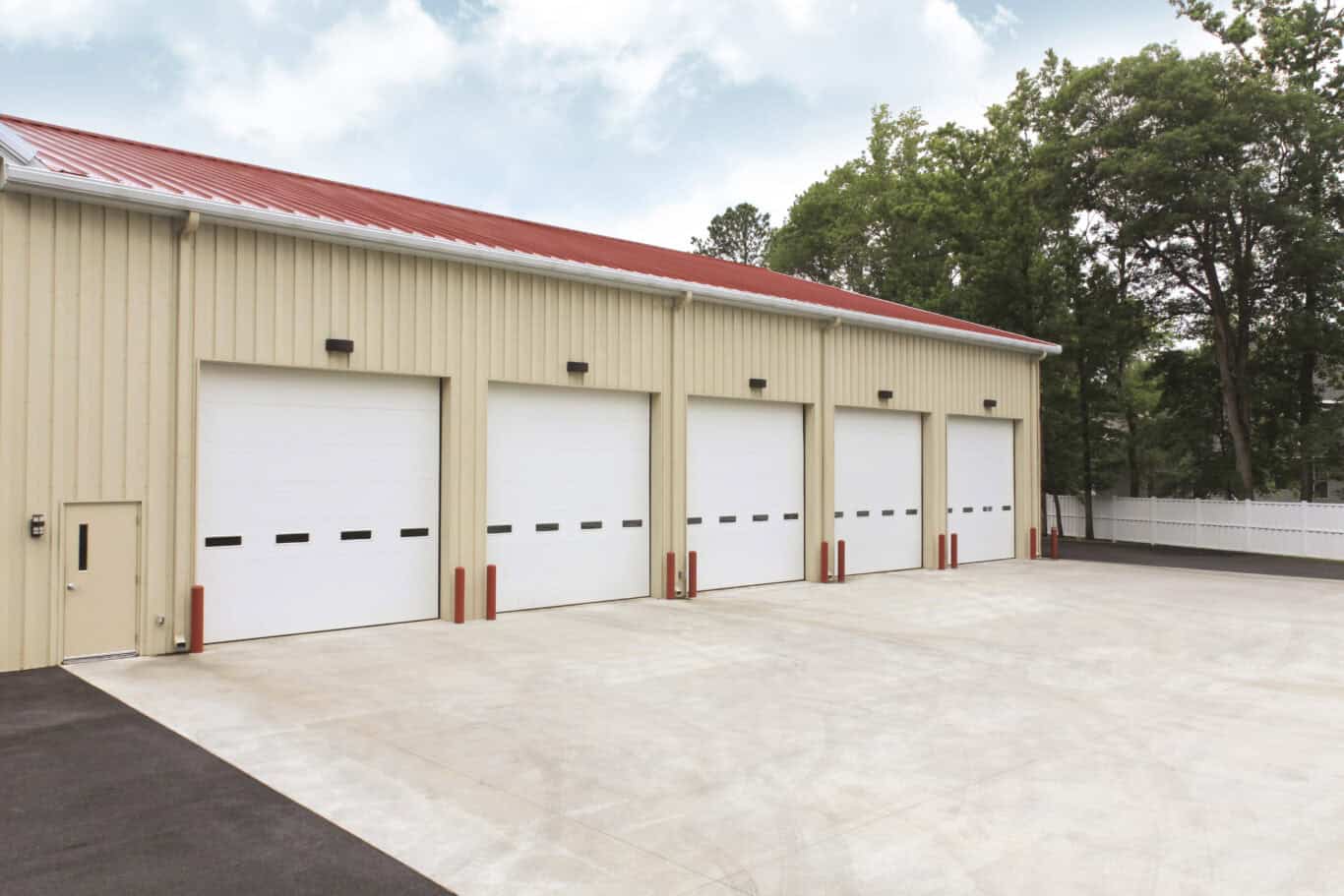 A beige warehouse with four residential garage doors, each framed by red bollards, sits on a spacious concrete lot. Trees and a white fence enhance the backdrop.