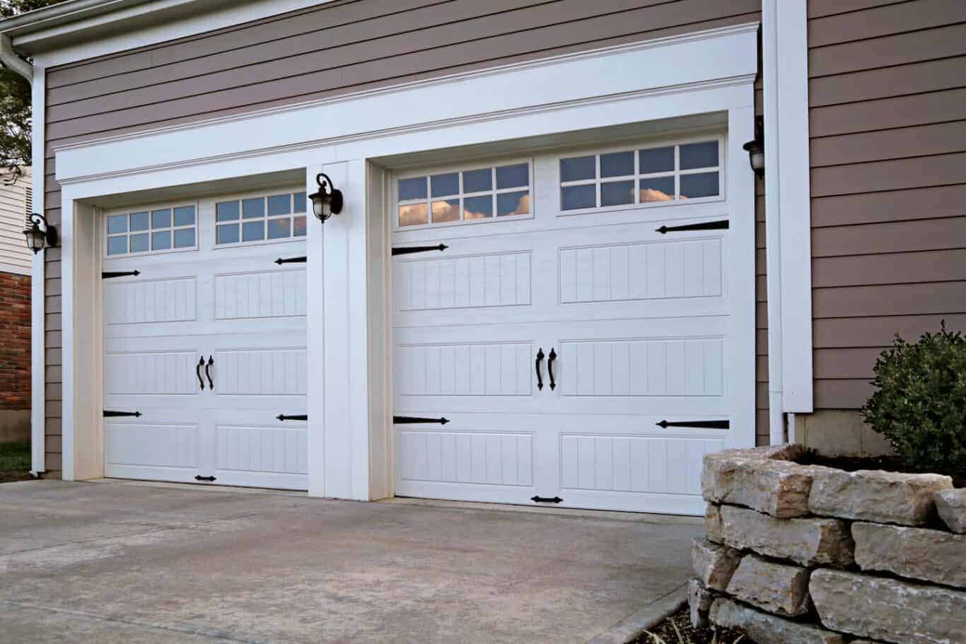 Two white garage doors with decorative windows and black handles on a gray house; stone planter and shrubs are visible on the right side.